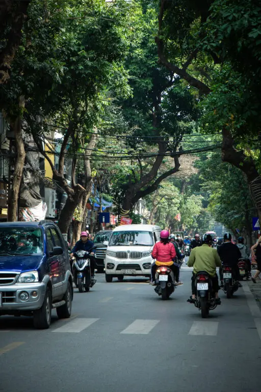 Straßenverkehr in Hanoi mit Rollern und schattigen Bäumen