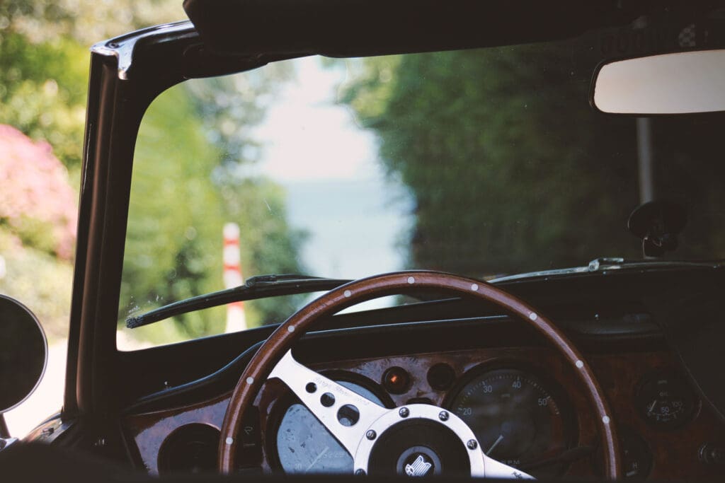 Fotografie durch ein Oldtimer-Lenkrad mit Baumallee und Meer im Hintergrund