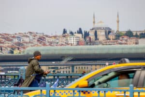 A fisherman casts his line from a bridge while a yellow taxi passes behind him, with the Istanbul skyline and mosque visible in the distance.