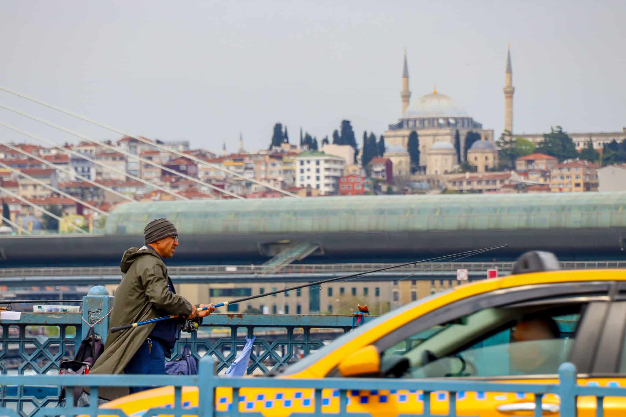 A fisherman casts his line from a bridge while a yellow taxi passes behind him, with the Istanbul skyline and mosque visible in the distance.