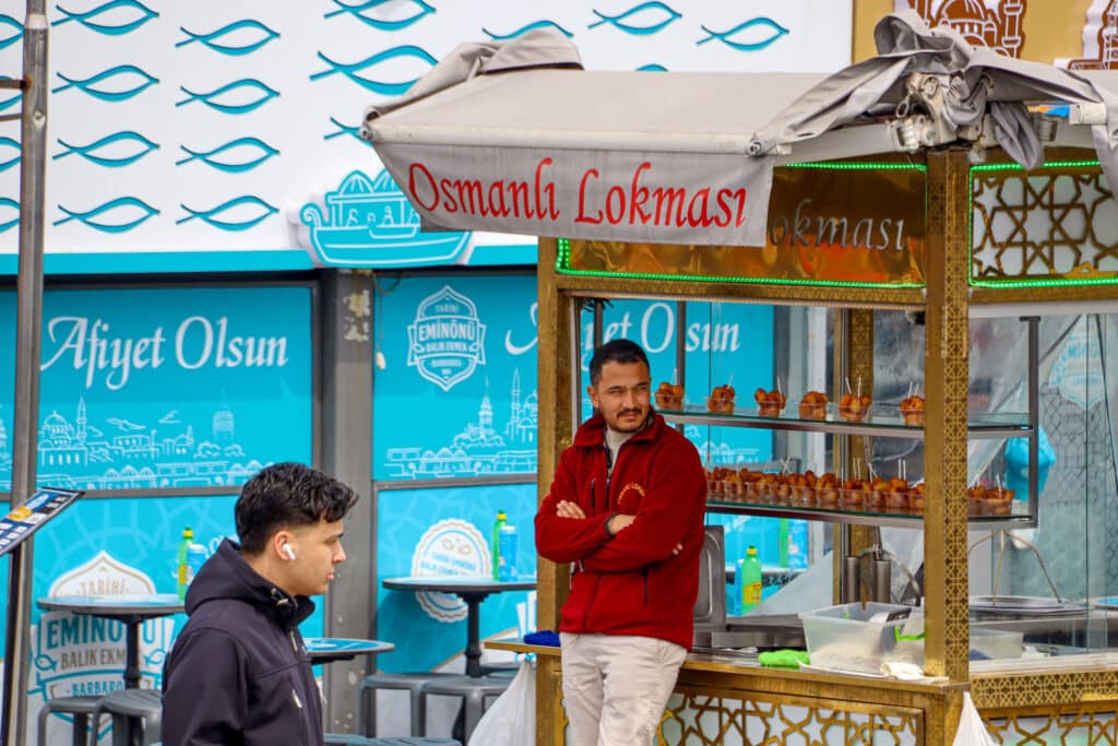 A vendor in traditional red costume stands at a colorful fish restaurant sign in a street market scene in Istanbul, Turkey