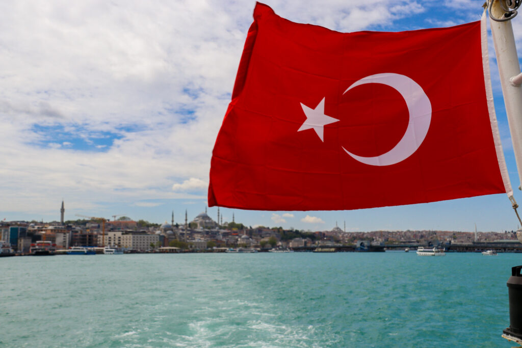 A large red Turkish flag with white crescent and star billows dramatically in the wind over the turquoise Bosphorus waters, Istanbul, Turkey.