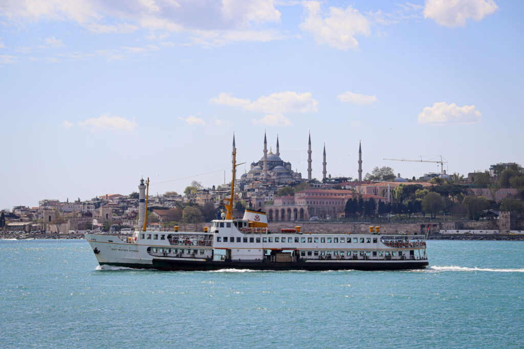 A passenger ferry crosses the Bosphorus with the Istanbul skyline and Blue Mosque minarets visible in the background under a blue sky.