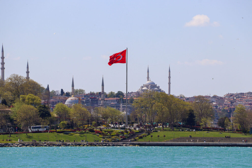 The Blue Mosque rises above a green tree-lined hillside with a Turkish flag in the foreground and the Bosphorus in front, Istanbul, Turkey.