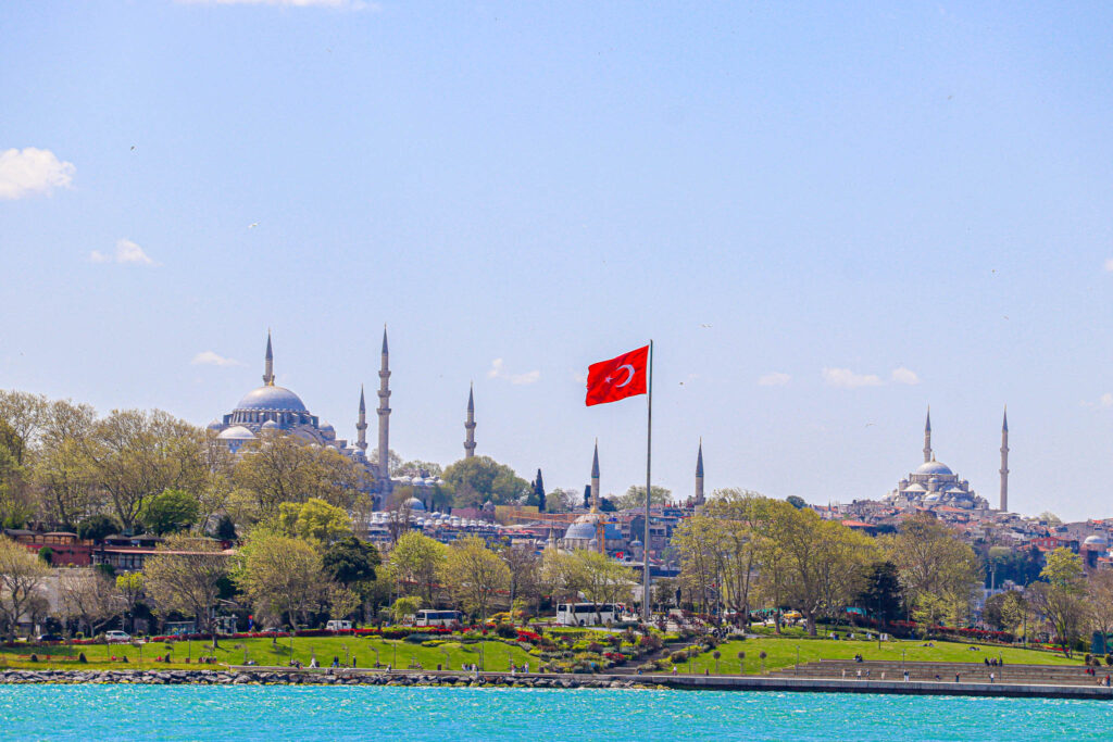 Wide panoramic view of the Istanbul skyline featuring multiple mosques and minarets with a Turkish flag in the foreground, seen from the Bosphorus.