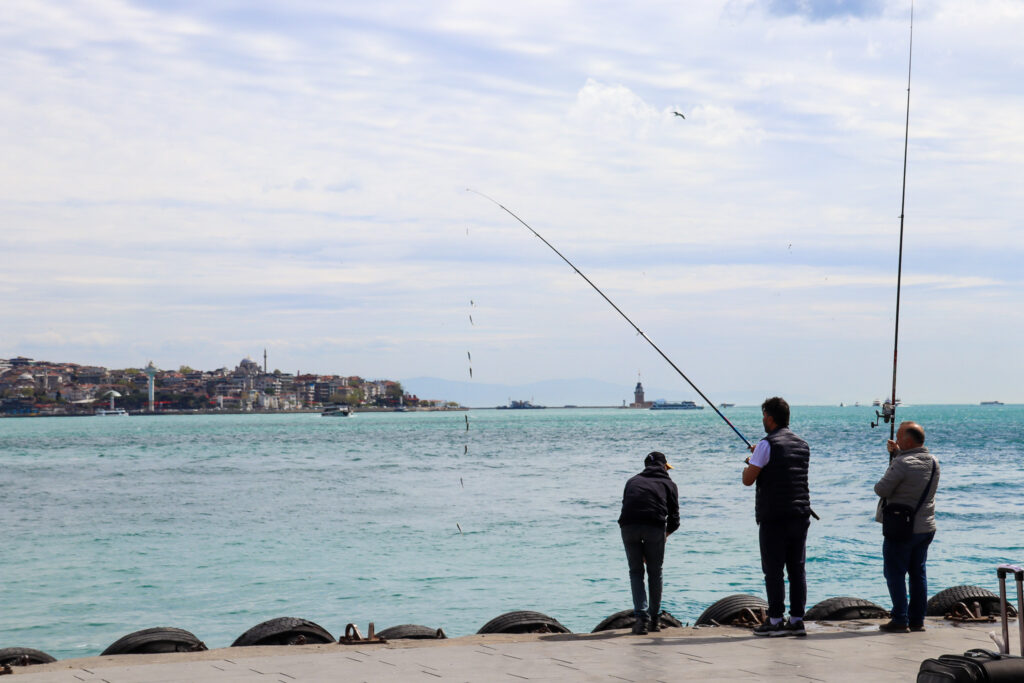 Two men fish from rocky shores with the iconic Maiden's Tower (Kız Kulesi) visible in the misty Bosphorus background, Istanbul, Turkey.