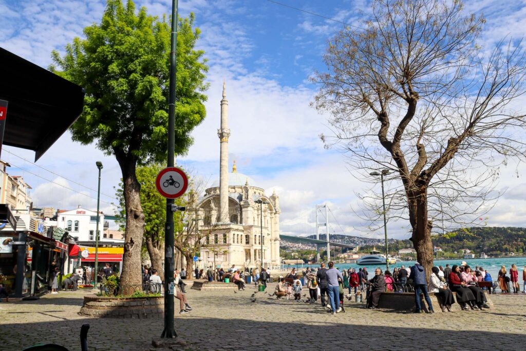 The Ortakoy Mosque square with a large bare tree, outdoor cafes and people strolling along the Bosphorus waterfront in Istanbul, Turkey.