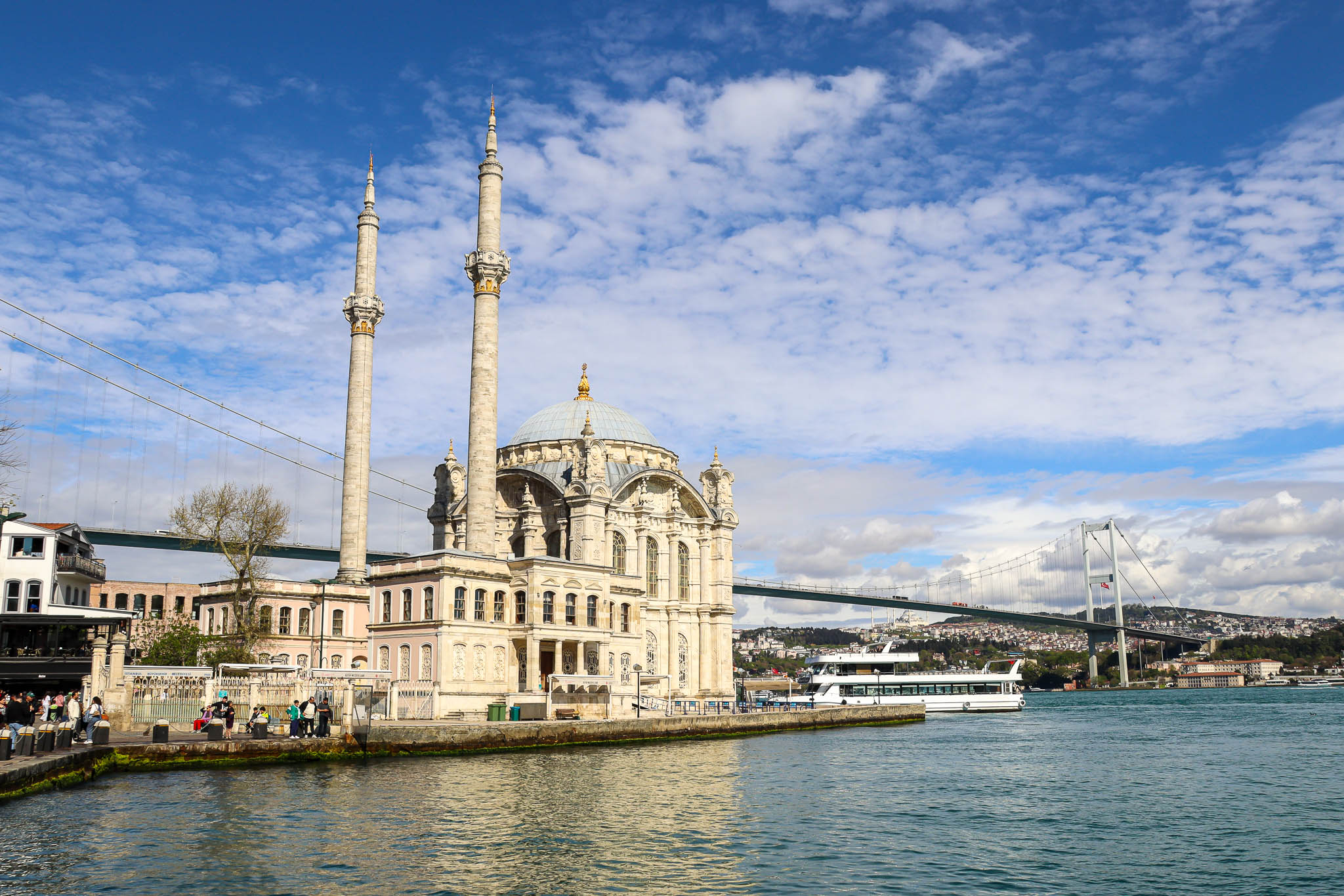 The iconic Ortakoy Mosque sits in the foreground with the massive Bosphorus Bridge (15 July Martyrs Bridge) spanning behind it on a sunny day, Istanbul.