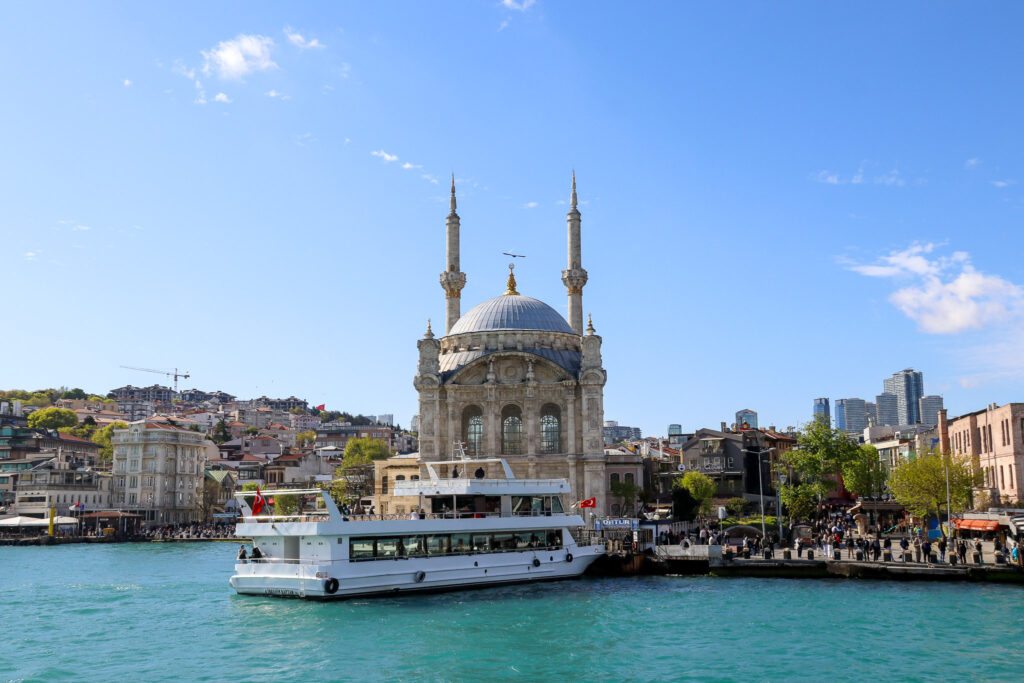 The Ortakoy Mosque is seen alongside a passing passenger ferry with the Bosphorus Bridge visible above on a sunny day in Istanbul, Turkey.