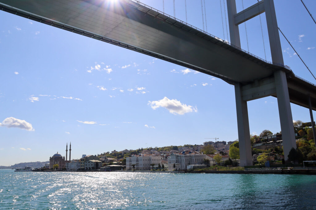 Dramatic perspective looking up and along the Bosphorus Bridge from below, with sunlight streaming through the structure and reflecting on the water, Istanbul.