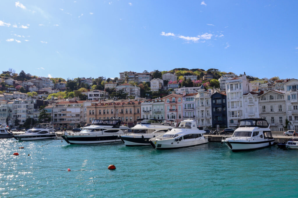 A busy marina with numerous yachts and boats moored in the foreground with the Istanbul mosque skyline stretching across the background on a sunny day.