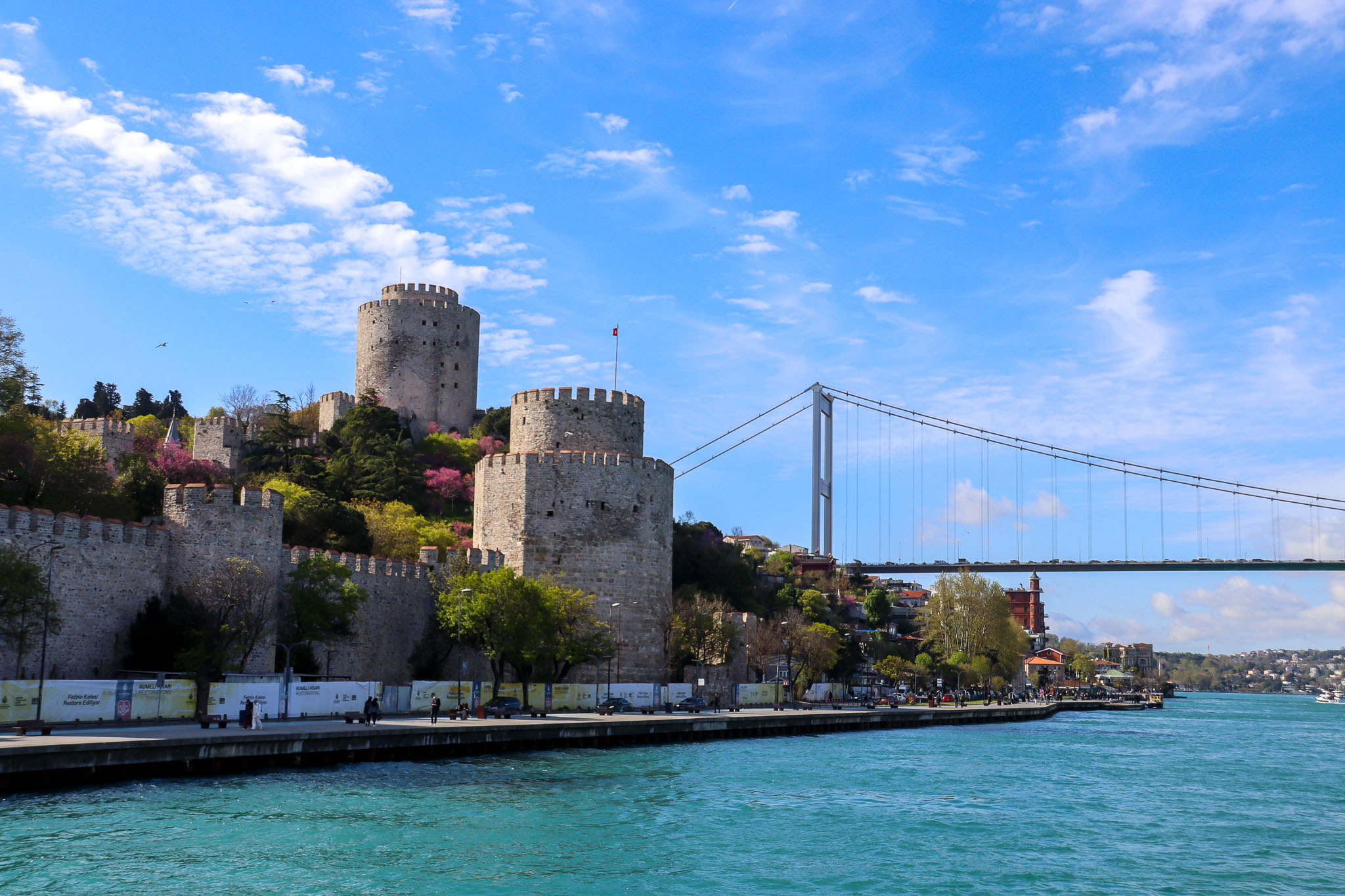 Wide view of the Rumeli Hisarı fortress with its multiple towers along the Bosphorus shoreline and the suspension bridge spanning the strait behind it, Istanbul.
