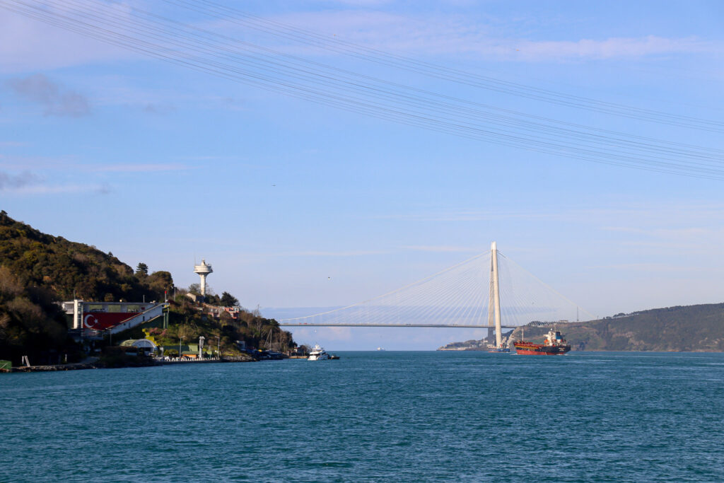 Wide view along the Bosphorus strait showing lush green hillside shores with the suspension bridge spanning the waterway on a sunny day, Istanbul