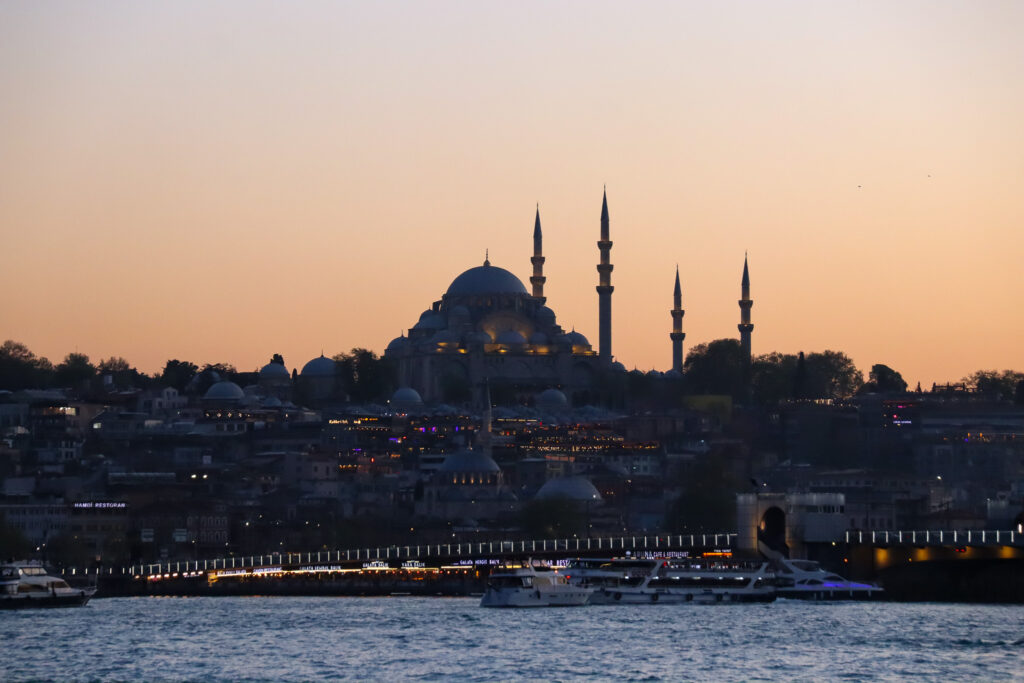 The Süleymaniye Mosque with its four minarets is dramatically silhouetted against a vivid orange and golden sunset sky over the Bosphorus, Istanbul, Turkey.