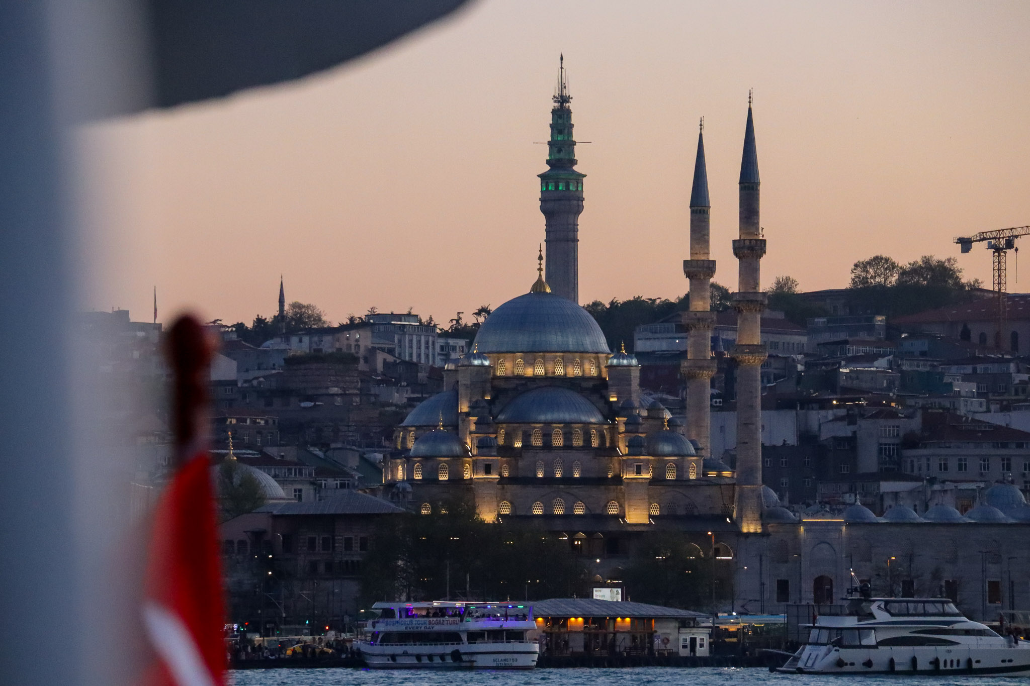 A mosque silhouette at golden sunset seen through a ferry window frame with a Turkish flag in the foreground, creating a layered composition, Istanbul.