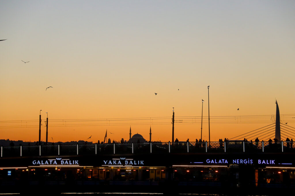 Wide panoramic silhouette of the entire Istanbul skyline with mosque minarets against a brilliant deep orange sunset sky, approaching Galata Bridge, Turkey.