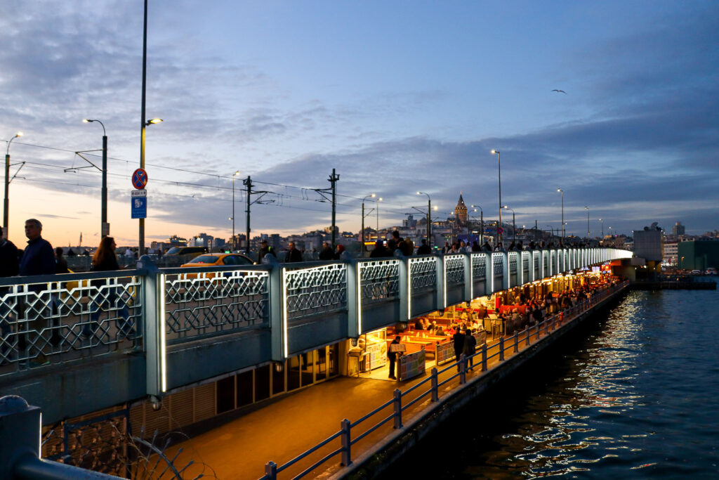 The Galata Bridge glows with warm golden lights at blue hour dusk with fishermen lining the railings and the Istanbul skyline softly lit in the background, Turkey.